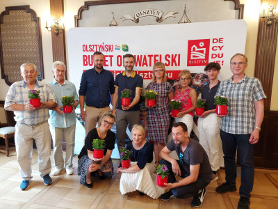 grupa ludzi na tle planszy z napisem Olsztyński budżet Obywatelski, wszyscy trzymają czerwone doniczki z kwiatkami w dłoniach.