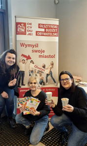 Na pierwszym planie trzy kobiety z kubkami w rękach na tle tablicy informacyjnej z hasłem: wymyśl swoje miasto. 